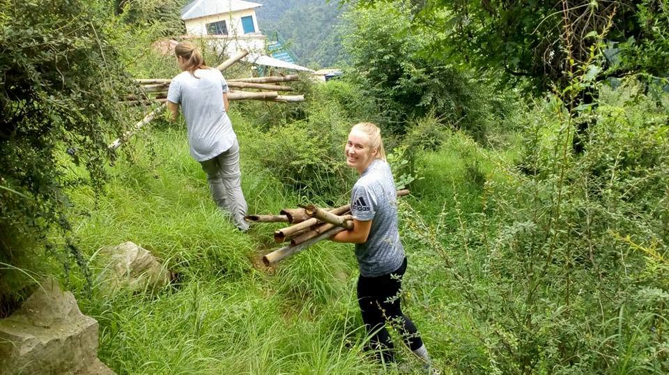 A volunteer carrying bamboo sticks, looking behind at the camera, as another volunteer carrying bamboo sticks leads on in the background.